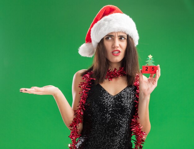 Lado confundido joven hermosa chica con sombrero de Navidad con guirnalda en el cuello sosteniendo juguete de Navidad mano extendida aislada en la pared verde