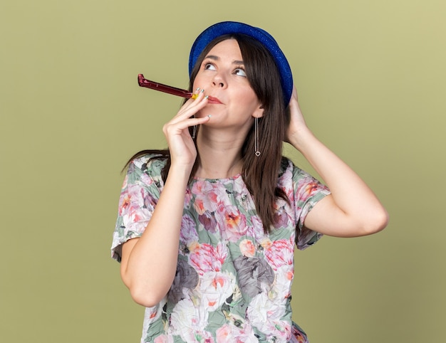 Lado de mirada alegre joven hermosa chica vistiendo gorro de fiesta soplando silbato de fiesta poniendo la mano en la cabeza