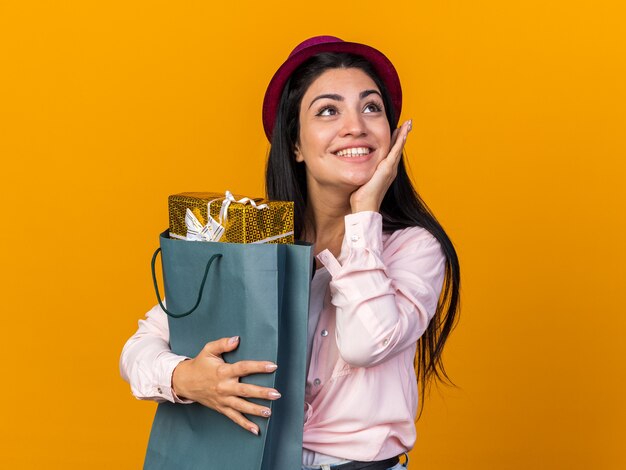 Lado de mirada complacido hermosa joven vistiendo gorro de fiesta sosteniendo una bolsa de regalo poniendo la mano en la mejilla