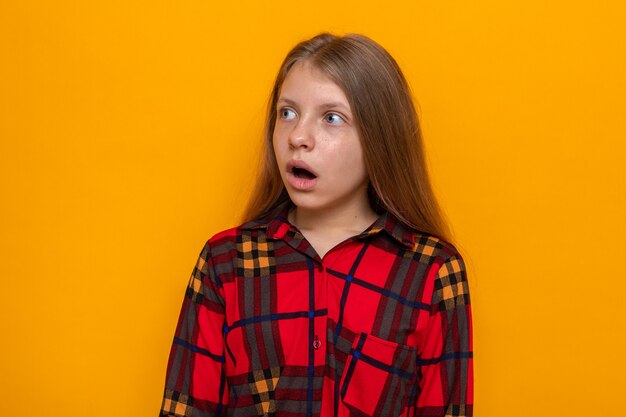 Lado mirando asustado hermosa niña con camisa roja aislada en la pared naranja