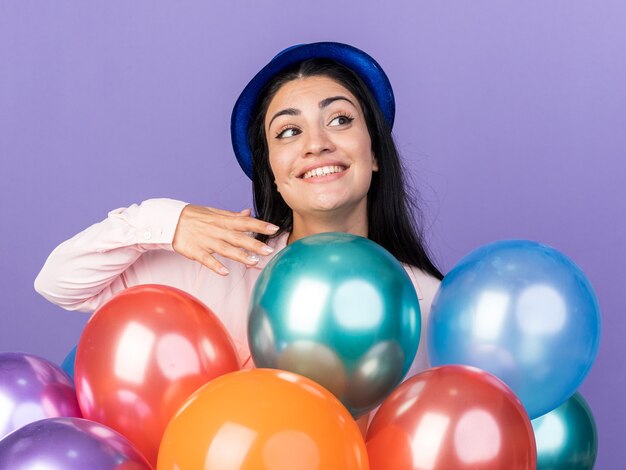 Foto gratuita lado mirando sonriente joven hermosa chica con gorro de fiesta de pie detrás de globos