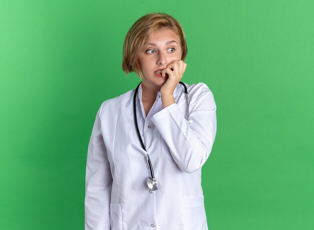 Lado preocupado joven doctora vistiendo bata médica con estetoscopio muerde las uñas aisladas en la pared verde