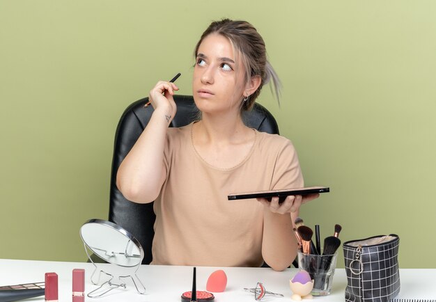 Lado sorprendido joven hermosa niña sentada en la mesa con herramientas de maquillaje sosteniendo pincel con paleta de sombra de ojos aislado sobre fondo verde oliva