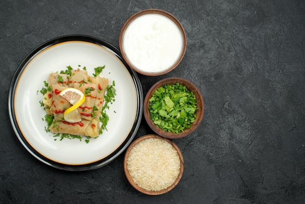 Lado superior del lado apetitoso repollo relleno con hierbas de limón y salsa en un plato blanco y hierbas de arroz y crema agria en tazones sobre mesa negra