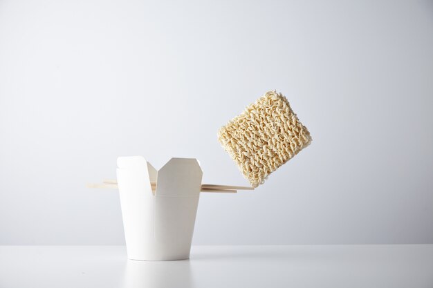 Ladrillo de fideos secos se equilibra en el borde de los palillos en la caja de comida para llevar en blanco aislada en el conjunto minorista comercial blanco