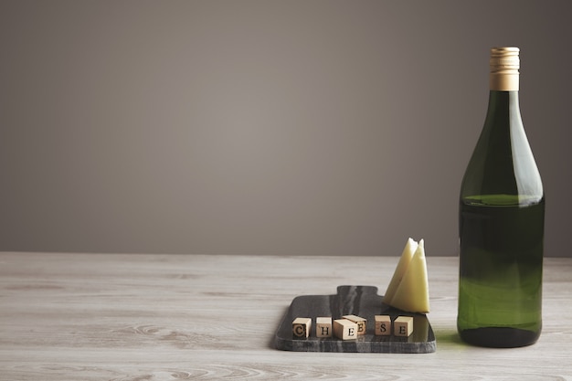 Ladrillo de letra de madera cerca de queso de cabra en rodajas y botella de uva de jugo de vino verde medio vacía aislada en tablero de piedra de mármol sobre mesa de madera blanca y fondo gris neutro