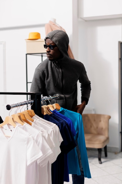 Foto gratuita ladrón afroamericano que elige robar mercancía de moda, mirando a su alrededor para ver si alguien lo está observando. ladrón robando una boutique moderna, con capucha y gafas de sol. concepto de crimen