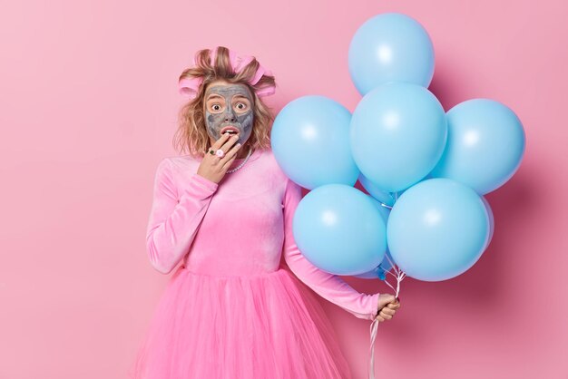 Ladyy sorprendida con máscara facial aplicada y rodillos para el cabello se prepara para ocasiones especiales con vestidos festivos que sostienen un montón de globos azules aislados sobre fondo rosa Dios mío, no estoy listo para la fiesta