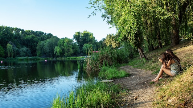 Un lago con muchos árboles verdes reflejados en el agua, dos niñas están sentadas en la orilla y cañas a lo largo de ella en Chisinau, Moldavia