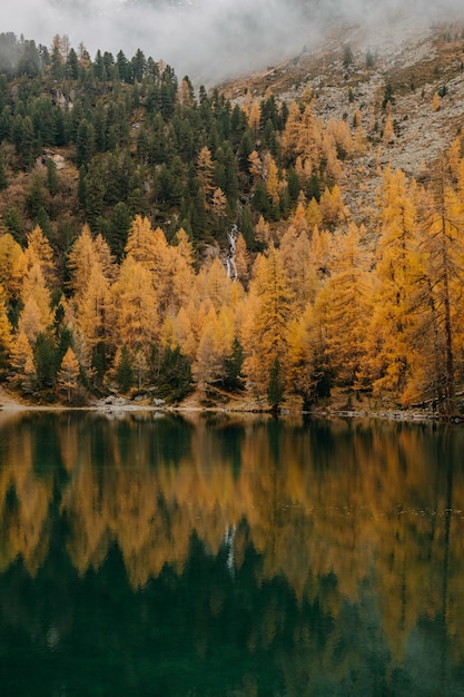 Lago tranquilo y nubes bajas que cubren una montaña áspera cubierta con follaje otoñal colorido