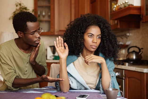 Lamentable culpable joven afroamericano con gafas que ofrece la mano a su novia enojada en señal de reconciliación después de una pelea seria, pero la mujer parece rechazar todas las disculpas y excusas