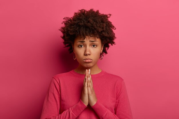 La lamentable mujer miserable con peinado afro junta las palmas de las manos, pide favor, tiene una expresión facial triste, tímida e inocente, viste un suéter rosa