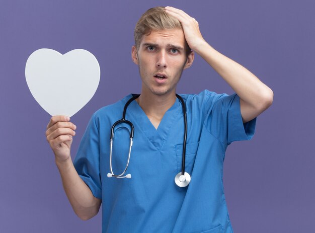 Lamentado joven médico vistiendo uniforme médico con estetoscopio sosteniendo una caja en forma de corazón poniendo la mano en la cabeza aislada en la pared azul