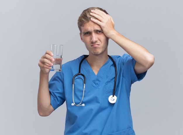 Lamentado joven médico vistiendo uniforme médico con estetoscopio sosteniendo un vaso de agua poniendo la mano en la cabeza aislada en la pared blanca