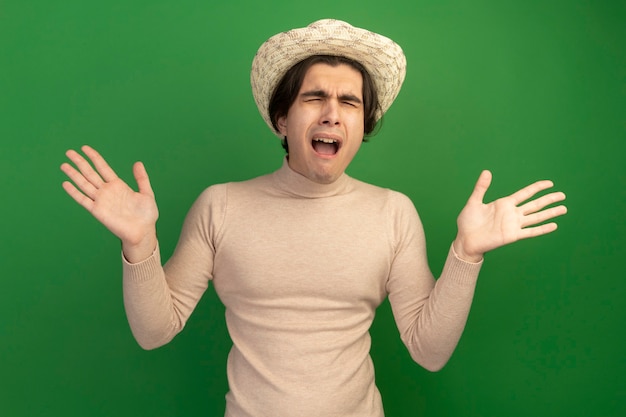 Lamentado con los ojos cerrados joven guapo con sombrero extendiendo las manos aisladas en la pared verde