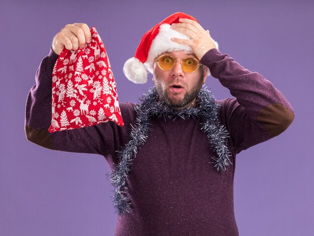 Lamentando al hombre de mediana edad con gorro de Papá Noel y guirnalda de oropel alrededor del cuello con gafas sosteniendo un saco de regalo de Navidad manteniendo la mano en la cabeza aislada en la pared púrpura