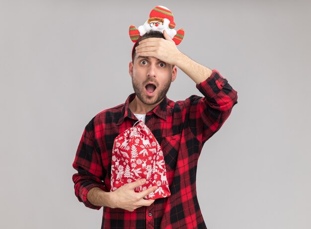 Lamentando a joven caucásico con diadema de navidad sosteniendo saco de navidad manteniendo la mano en la frente mirando a cámara aislada sobre fondo blanco