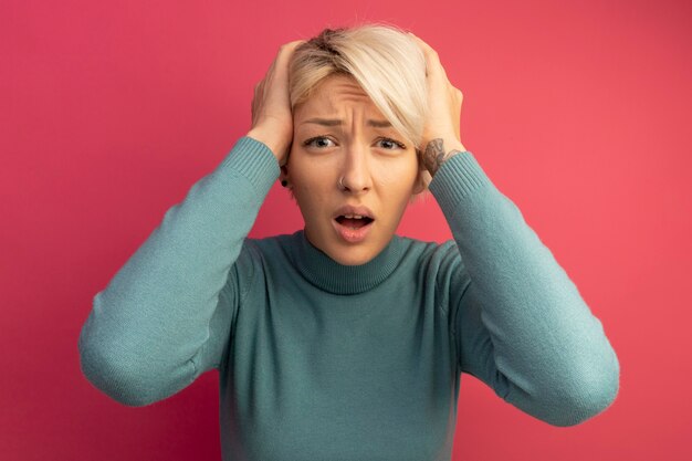 Lamentando a la joven rubia poniendo las manos en la cabeza mirando aislado en la pared rosa