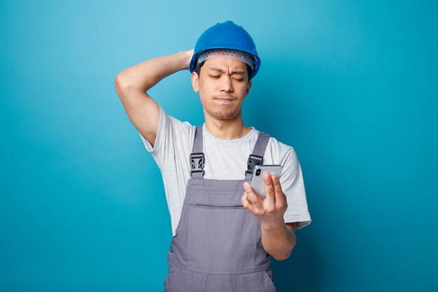 Lamentando a un joven trabajador de la construcción con casco de seguridad y uniforme manteniendo la mano detrás de la cabeza sosteniendo y mirando el teléfono móvil