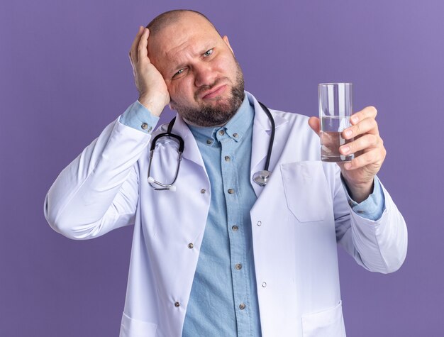 Lamentando a un médico varón de mediana edad con una bata médica y un estetoscopio sosteniendo un vaso de agua manteniendo la mano en la cabeza mirando al frente aislado en la pared púrpura