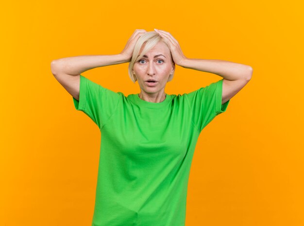 Lamentando la mujer eslava rubia de mediana edad mirando a la cámara manteniendo las manos en la cabeza aisladas sobre fondo amarillo