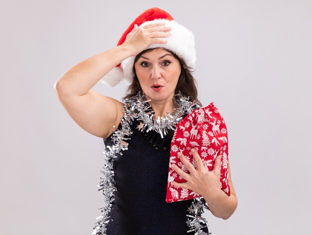 Lamentando a la mujer de mediana edad con gorro de Papá Noel y guirnalda de oropel alrededor del cuello sosteniendo un saco de regalo de Navidad mirando a la cámara manteniendo la mano en la cabeza aislada sobre fondo blanco con espacio de copia