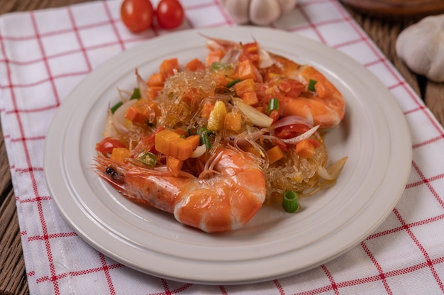 Langostinos salteados con fideos de vidrio en un plato blanco colocado sobre un paño con huevos y ajo.