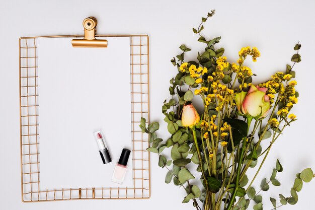 Lápiz labial; barniz de uñas en papel sobre el portapapeles dorado metálico con un ramo de flores aisladas sobre fondo blanco
