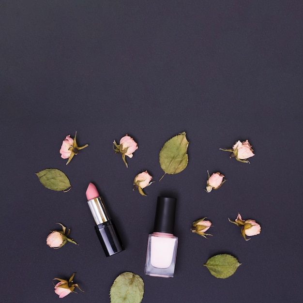 Lápiz labial rosado y botella de esmalte de uñas rodeado de brotes y hojas de rosa sobre fondo negro