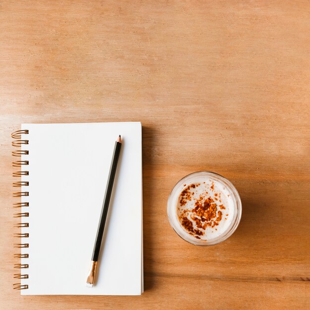 Lápiz sobre el cuaderno de espiral blanco cerrado y el vaso de café sobre fondo con textura de madera