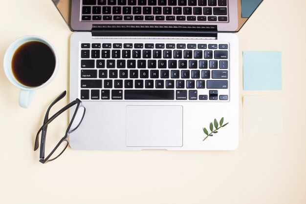 Una laptop abierta; taza de té gafas y bloc de notas sobre fondo beige