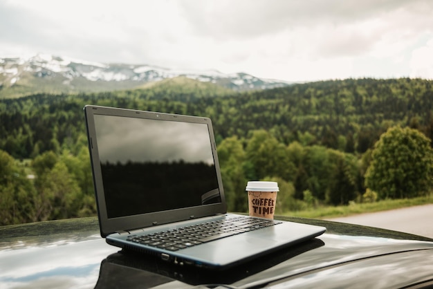 Laptop y un café para llevar en el capó del auto con hermosos paisajes naturales en el fondo