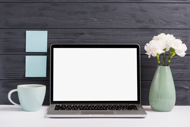 Laptop de pantalla blanca en blanco; florero; Copa en escritorio blanco contra pared de madera negra