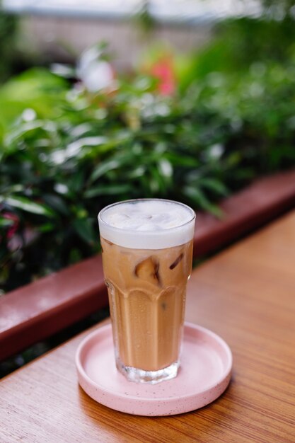 Latte helado en vidrio sobre un soporte rosa sobre una mesa de madera en el café de verano arbustos verdes