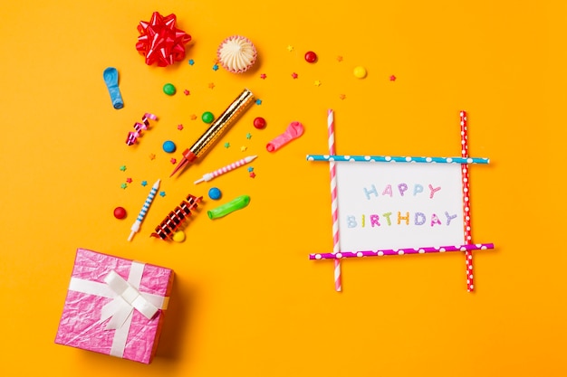 Lazo de cinta roja; aalaw; gemas serpentinas y salpicaduras de tarjeta de feliz cumpleaños y caja de regalo sobre fondo amarillo