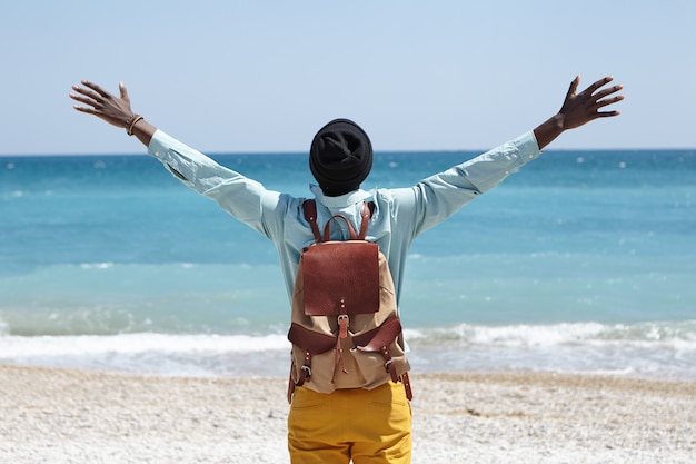 Lea la vista del feliz hombre afroamericano despreocupado de pie en la playa frente al mar azul, extendiendo los brazos, sintiendo libertad y conexión con la increíble naturaleza que lo rodea.