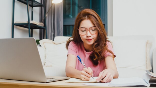 Lección de aprendizaje a distancia de joven estudiante adolescente de Asia con profesor en línea y estudio en computadora portátil en la sala de estar de casa por la noche. Distanciamiento social, cuarentena para la prevención del coronavirus.