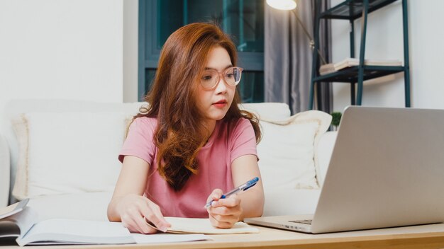 Lección de aprendizaje a distancia de joven estudiante adolescente de Asia con profesor en línea y estudio en computadora portátil en la sala de estar de casa por la noche. Distanciamiento social, cuarentena para la prevención del coronavirus.