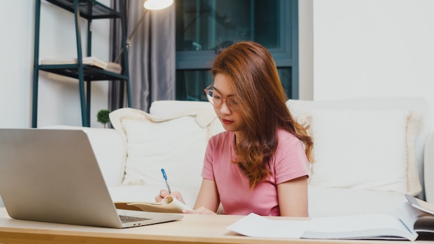 Lección de aprendizaje a distancia de joven estudiante adolescente de Asia con profesor en línea y estudio en computadora portátil en la sala de estar de casa por la noche. Distanciamiento social, cuarentena para la prevención del coronavirus.