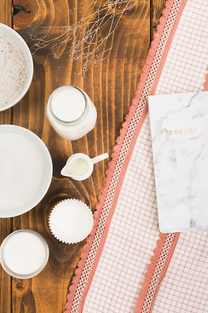 Leche; azúcar; Molde de harina y pastel con lista de tareas sobre tela texturizada sobre mesa con textura de madera