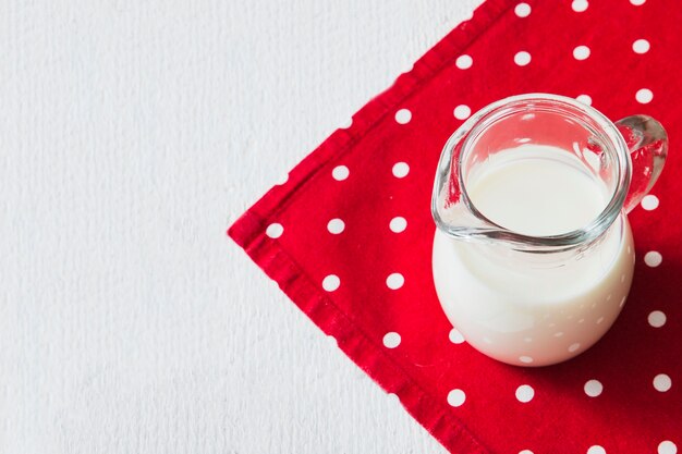 Leche en la jarra en la servilleta roja sobre el fondo blanco de madera