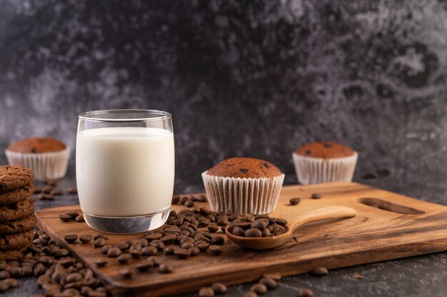 Leche en un vaso, completa con granos de café, pastelitos, plátanos y galletas en un plato de madera.