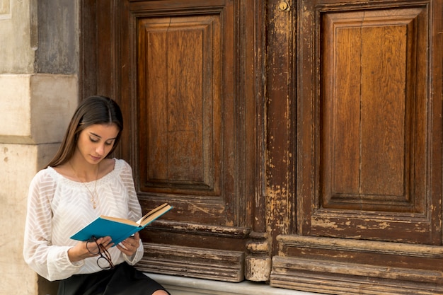 Foto gratuita lectura femenina joven mientras que se sienta cerca de puertas de madera