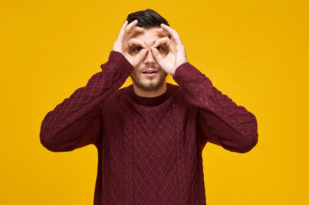 Lenguaje corporal y expresiones faciales humanas. Varón joven juguetón alegre en suéter que conecta los pulgares y los dedos índice, haciendo círculos alrededor de los ojos, mirando a través de agujeros como usando binoculares, espiando