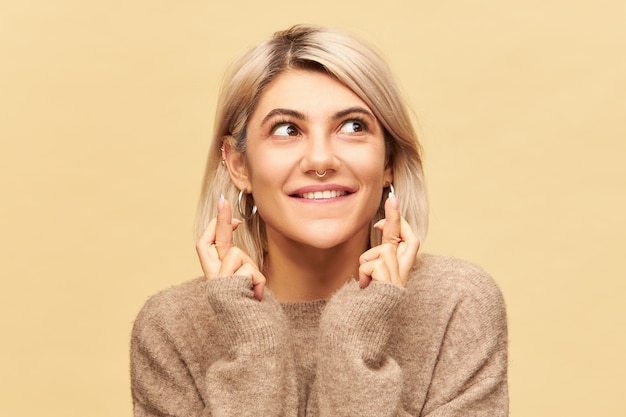 Lenguaje corporal. Retrato de adorable linda joven supersticiosa con cabello rubio y anillo en la nariz cruzando los dedos para tener buena suerte, deseando ganar en la lotería, con los ojos llenos de esperanza e impaciencia