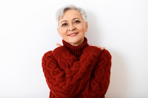 Lenguaje corporal. Retrato de una hermosa mujer europea mayor de moda calentándose en un día frío de invierno, cruzando los brazos sobre su pecho y sonriendo, vestida con un acogedor suéter de cuello alto burdeos