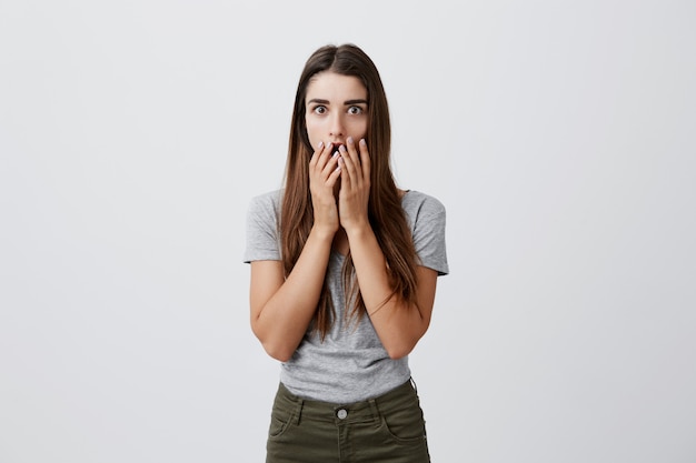 Lenguaje corporal. Retrato de joven atractiva chica caucásica encantadora con cabello largo y oscuro en camiseta gris informal y jeans ropa boca con las manos con las cejas levantadas