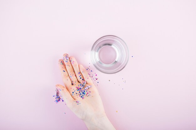 Lentejuelas y cápsula de glitter en mano humana cerca del vaso de agua sobre fondo rosa