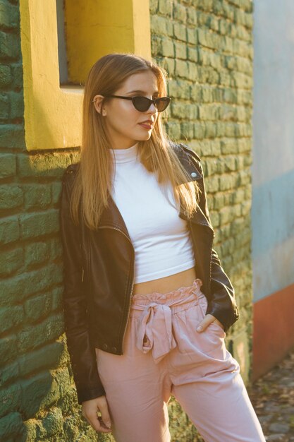 Lentes que llevan de moda hermosos de la mujer joven que se inclinan contra la pared de ladrillo verde