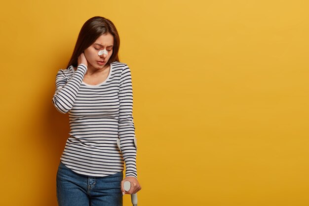 Lesiones en las piernas y restricción del movimiento humano. Mujer disgustada toca el cuello, siente dolor, se lesionó durante un viaje peligroso, tiene la nariz rota, posa contra la pared amarilla, espacio en blanco a un lado.